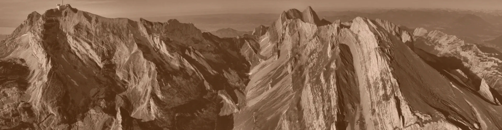 Tinted detail of a photo: Aerial view of steep limestone mountain ridges whose pale rock layers are tilted almost vertical, with green grassy slopes between them, a deep shadowed valley and a building with a mast on the left summit.