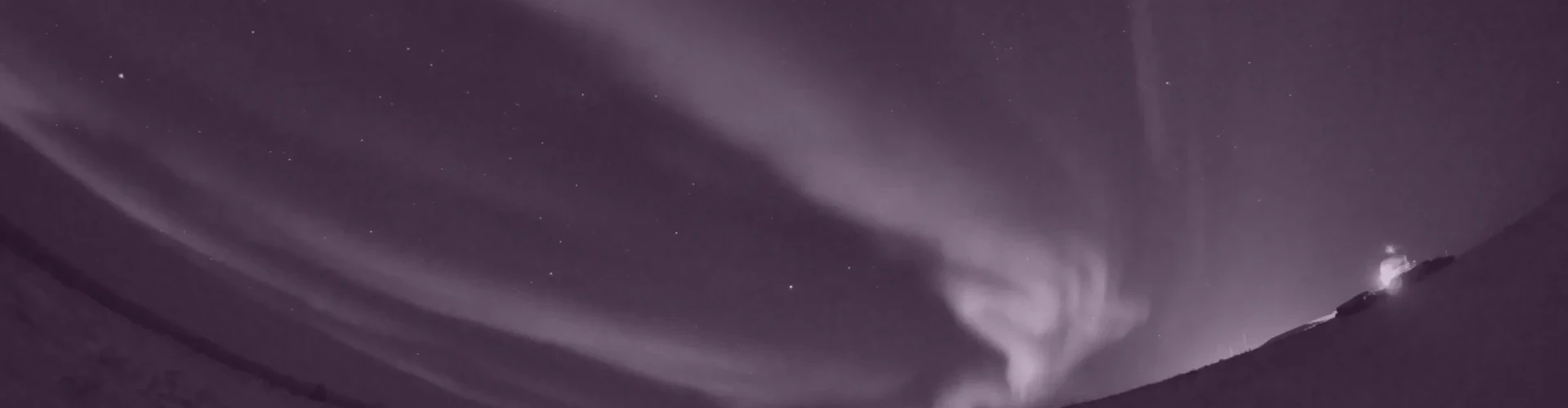 Tinted detail of a photo: Fisheye view of bright green aurora bands swirling across a starry night sky above a snowy curved horizon, with a small lit building at the right edge.