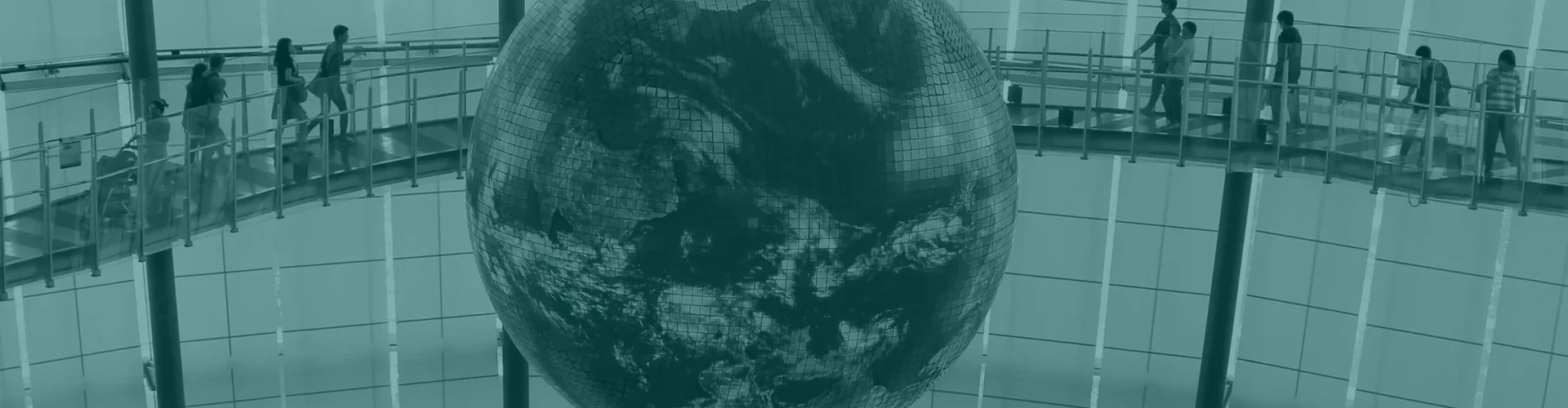 Tinted detail of a photo: Large glowing globe showing the Earth hanging in a museum atrium between two curved walkways with visitors, the surroundings in black and white and only the globe in colour.