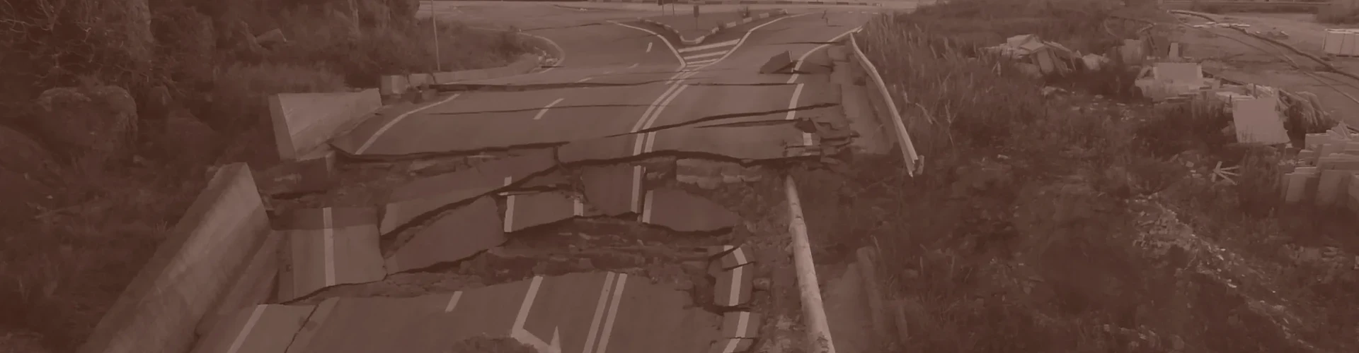Tinted detail of a photo: Road broken into slabs and buckled chunks of asphalt amid heaped soil, with a bent crash barrier and damaged buildings beside it, leading to a roundabout under a hazy setting sun.